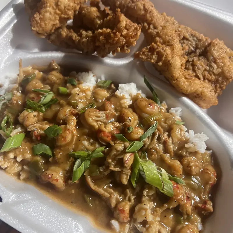Takeout container with crawfish tails in brown sauce over white rice with sliced green onion, next to golden fried pieces