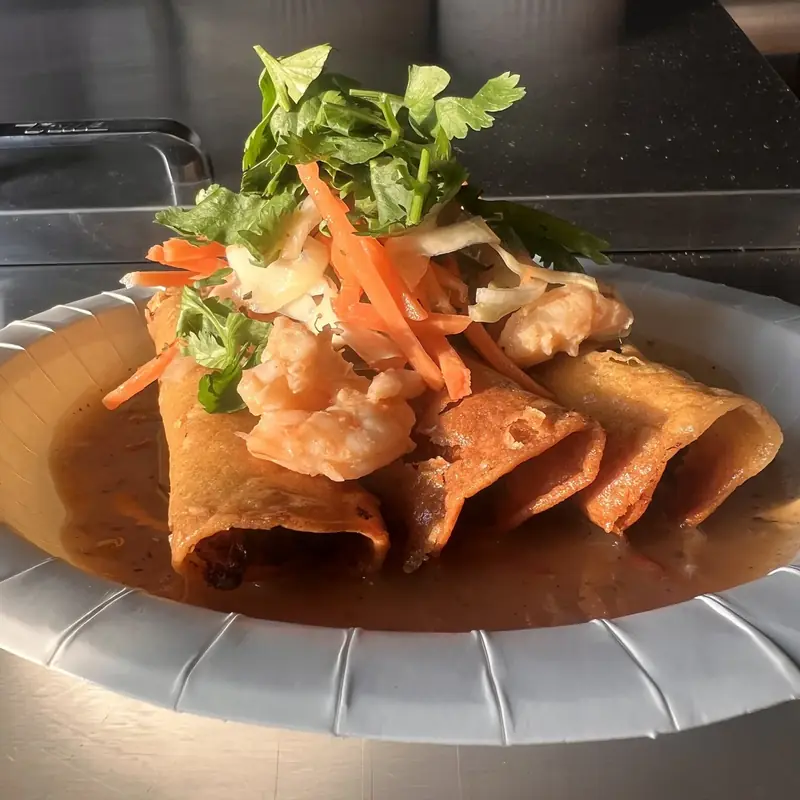 Rolled crisp tortillas in sauce on a paper plate, topped with shrimp, shredded carrot, sliced onion and leafy greens