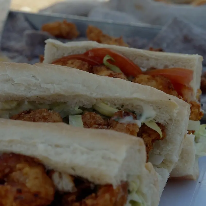 Sandwiches on long white rolls filled with fried pieces, shredded lettuce and tomato slices, lined up on a tray